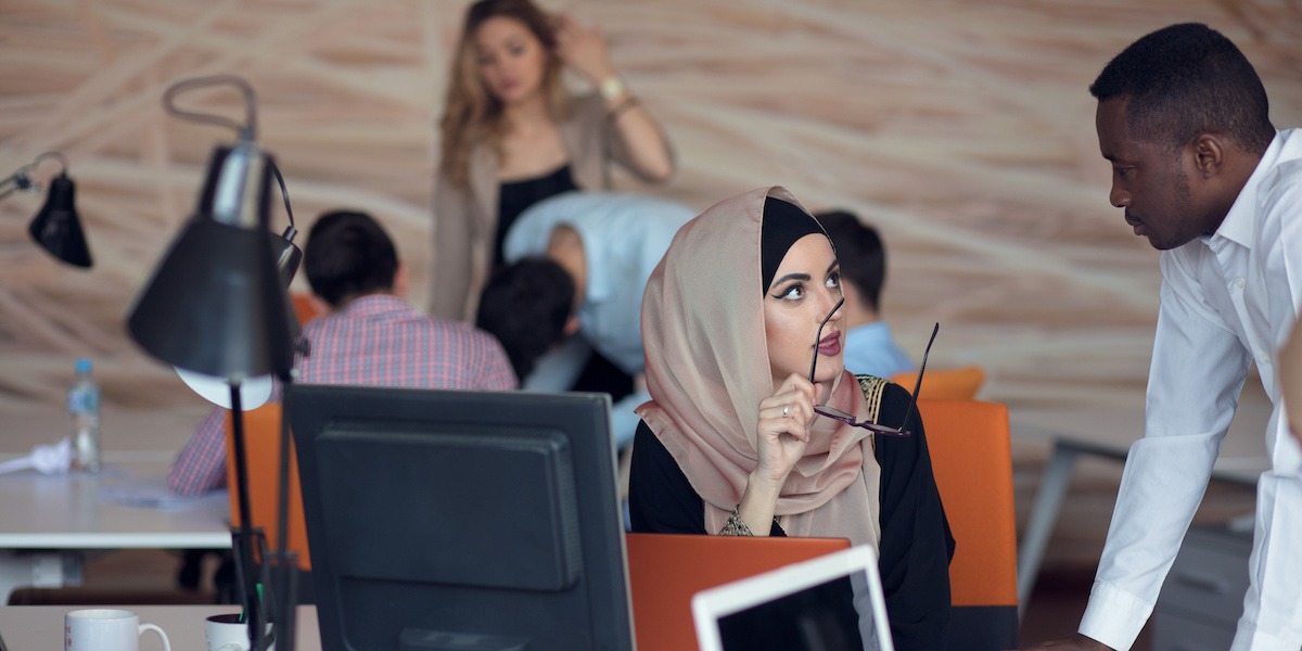 A product manager in a headscarf sits in a startup office explaining something to her colleague.